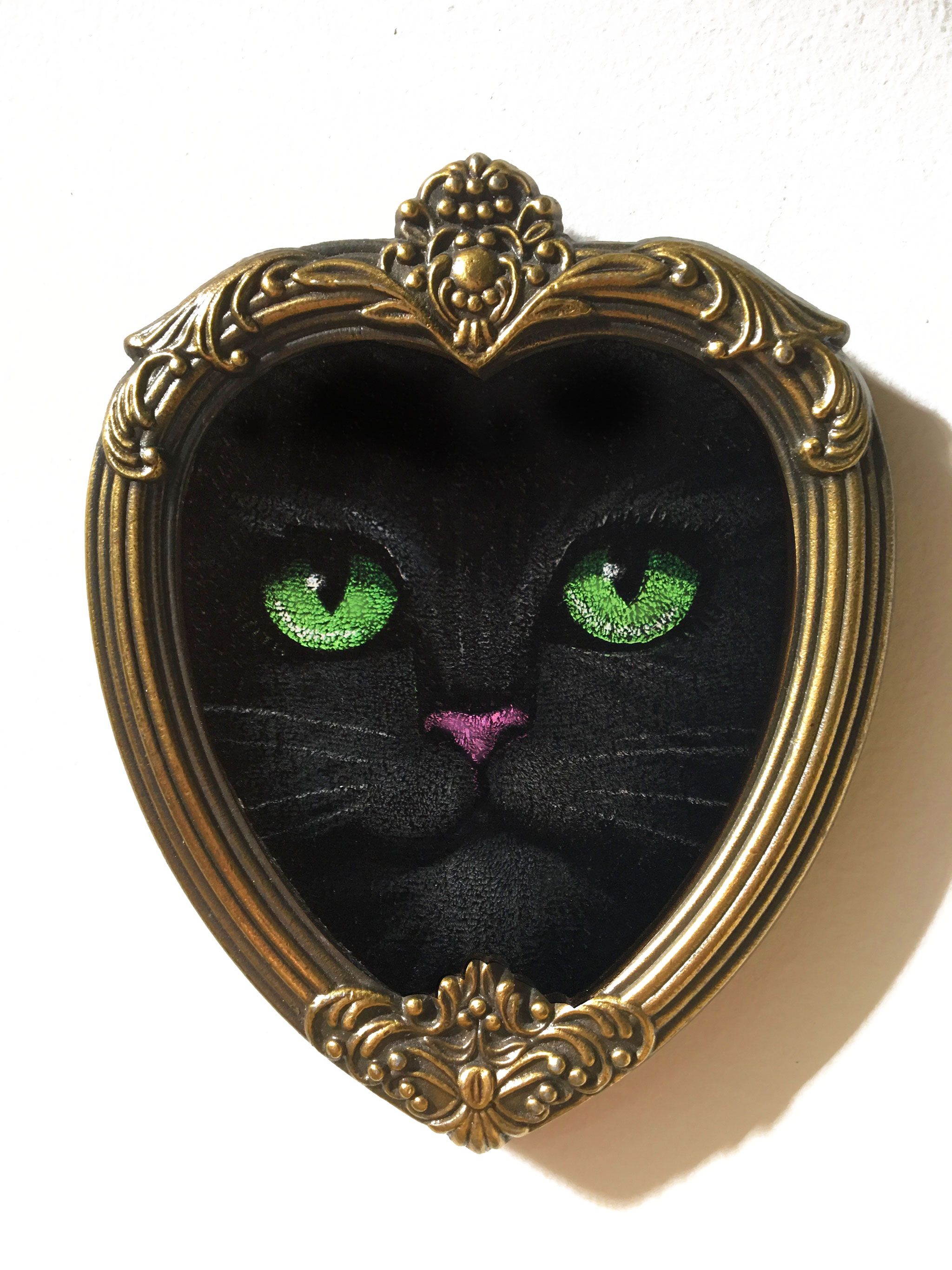 Heart Kitty on Velvet. Close-up of a black cat's face with bright green eyes and a pink nose, painted on black velvet in an ornate heart-shaped brass frame.