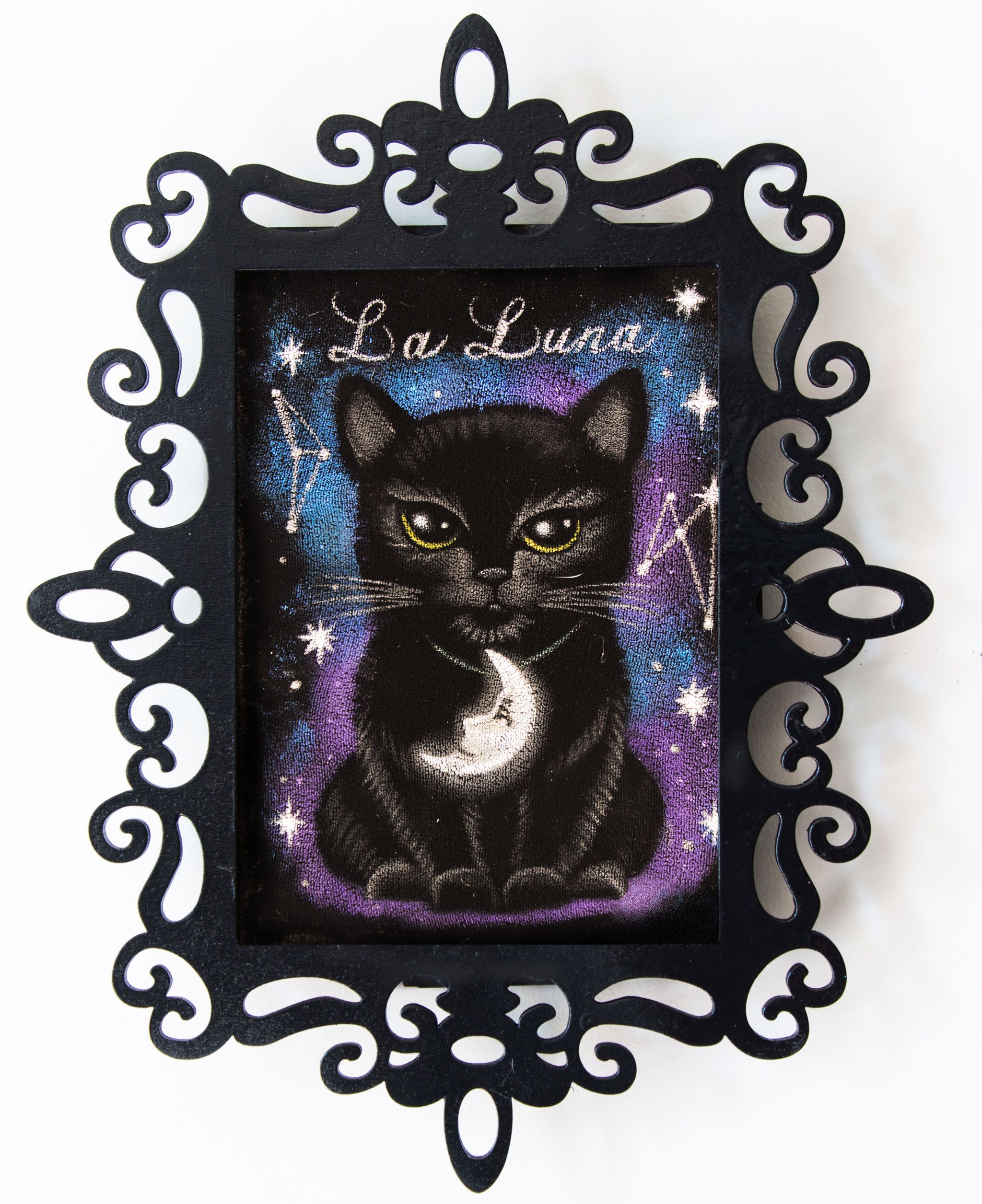La Luna Tarot Kitty On Velvet. A black kitten wearing a crescent moon pendant sits under the words La Luna, among stars and constellations in a blue and purple sky. Painted on black velvet in an ornate black filigree frame.